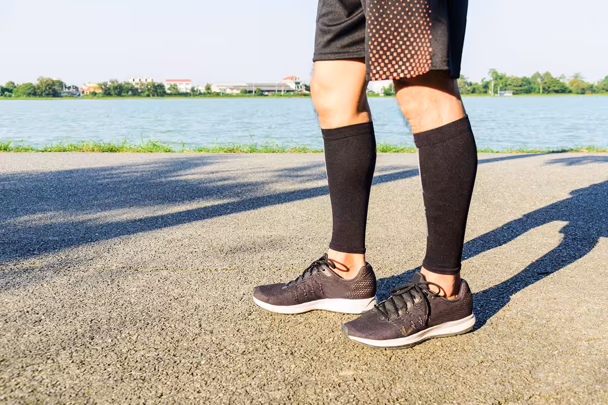 Person wearing black compression calf sleeves while standing outdoors on a paved path near a lake
