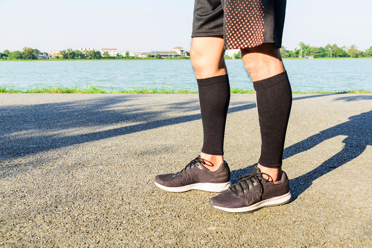 Person wearing black compression calf sleeves while standing outdoors on a paved path near a lake