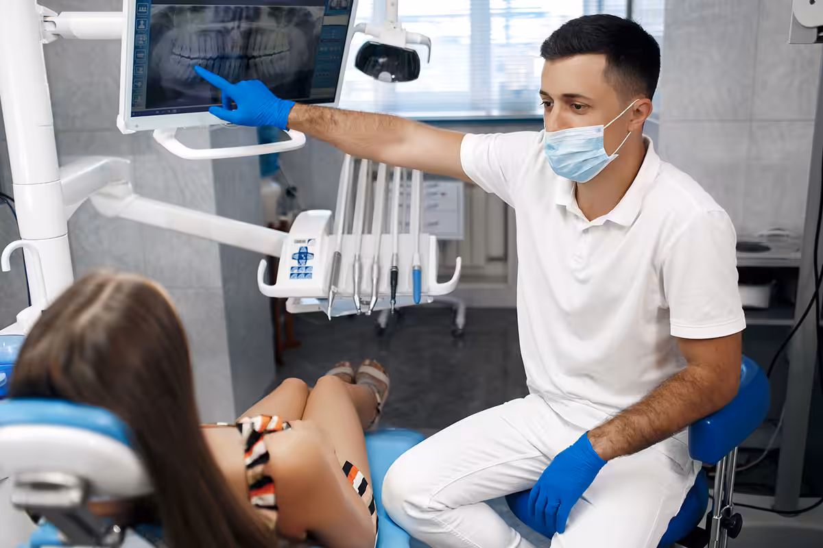 Dentist pointing to digital panoramic X-ray on monitor to explain orthodontic treatment options to reclined patient