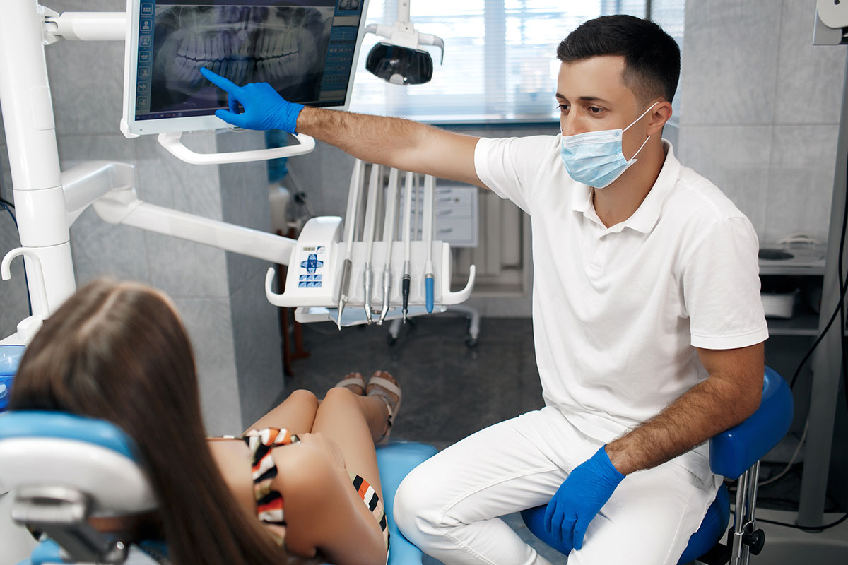 Dentist pointing to digital panoramic X-ray on monitor to explain orthodontic treatment options to reclined patient