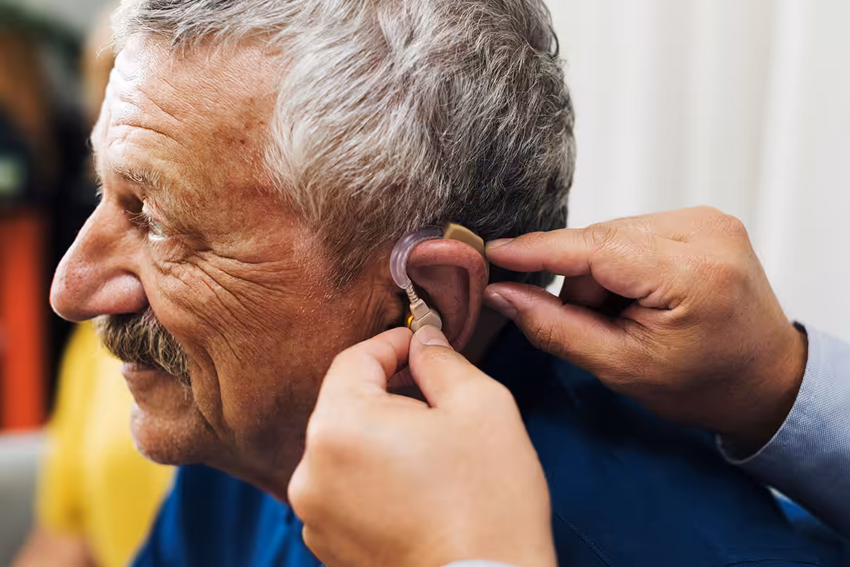 Audiologist fitting a behind-the-ear hearing aid on an older man covered under ASEBP MyRetiree Plan Extended Health Care benefits.