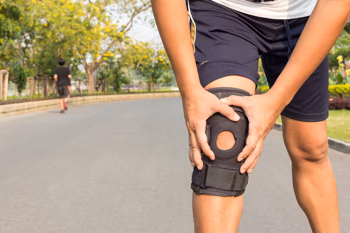 Person gripping knee fitted with black neoprene knee brace while exercising outdoors in a park setting
