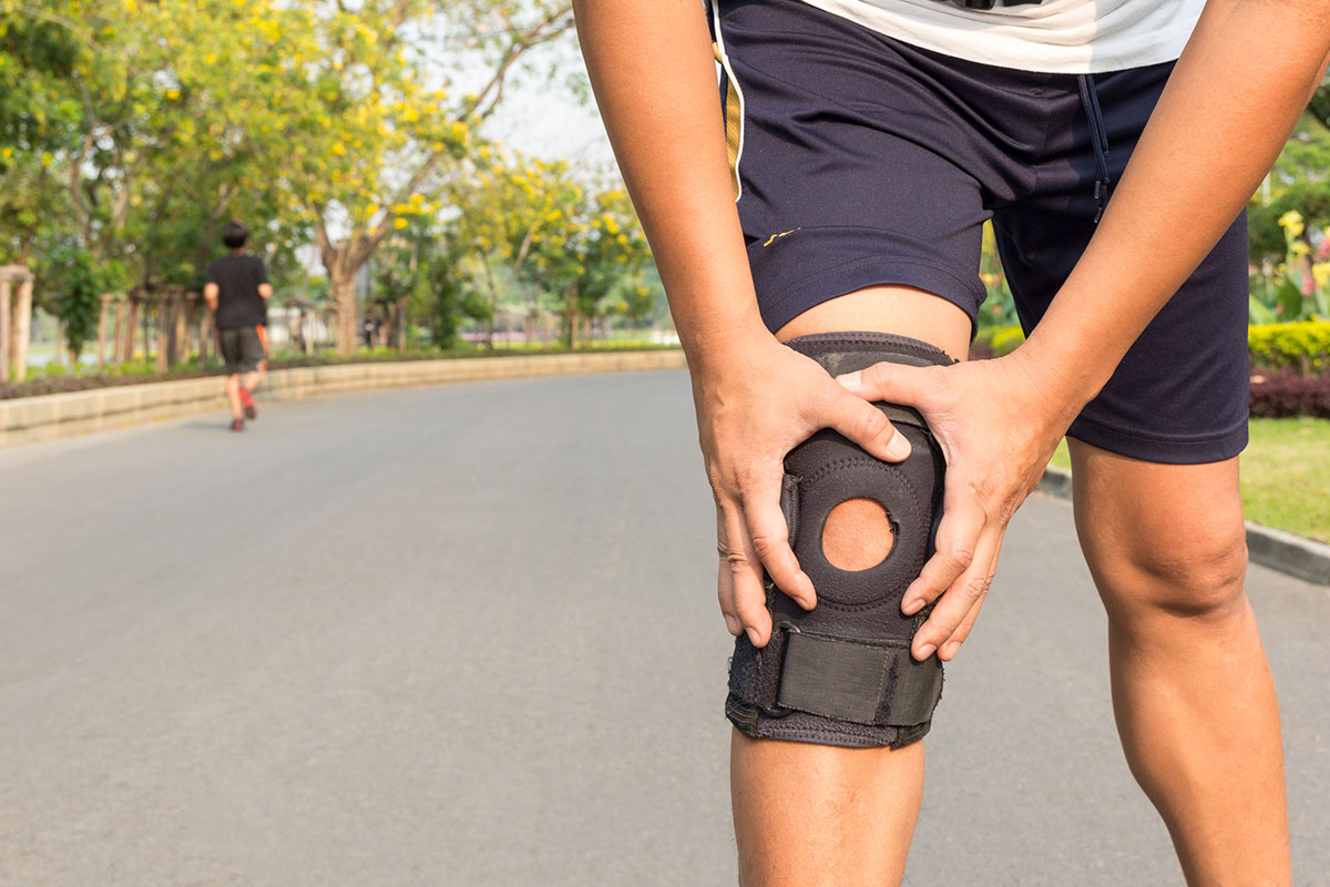 Person gripping knee fitted with black neoprene knee brace while exercising outdoors in a park setting