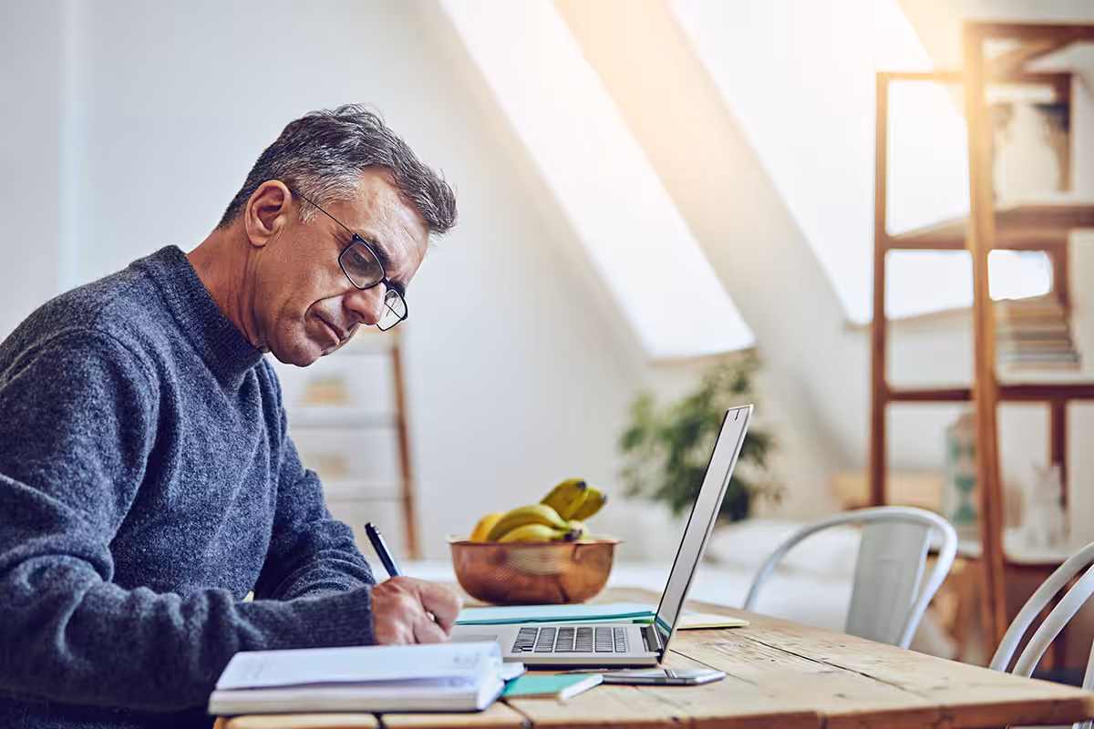 Retired Alberta educator carefully completing MyRetiree Plan premium payment paperwork at home desk with laptop nearby