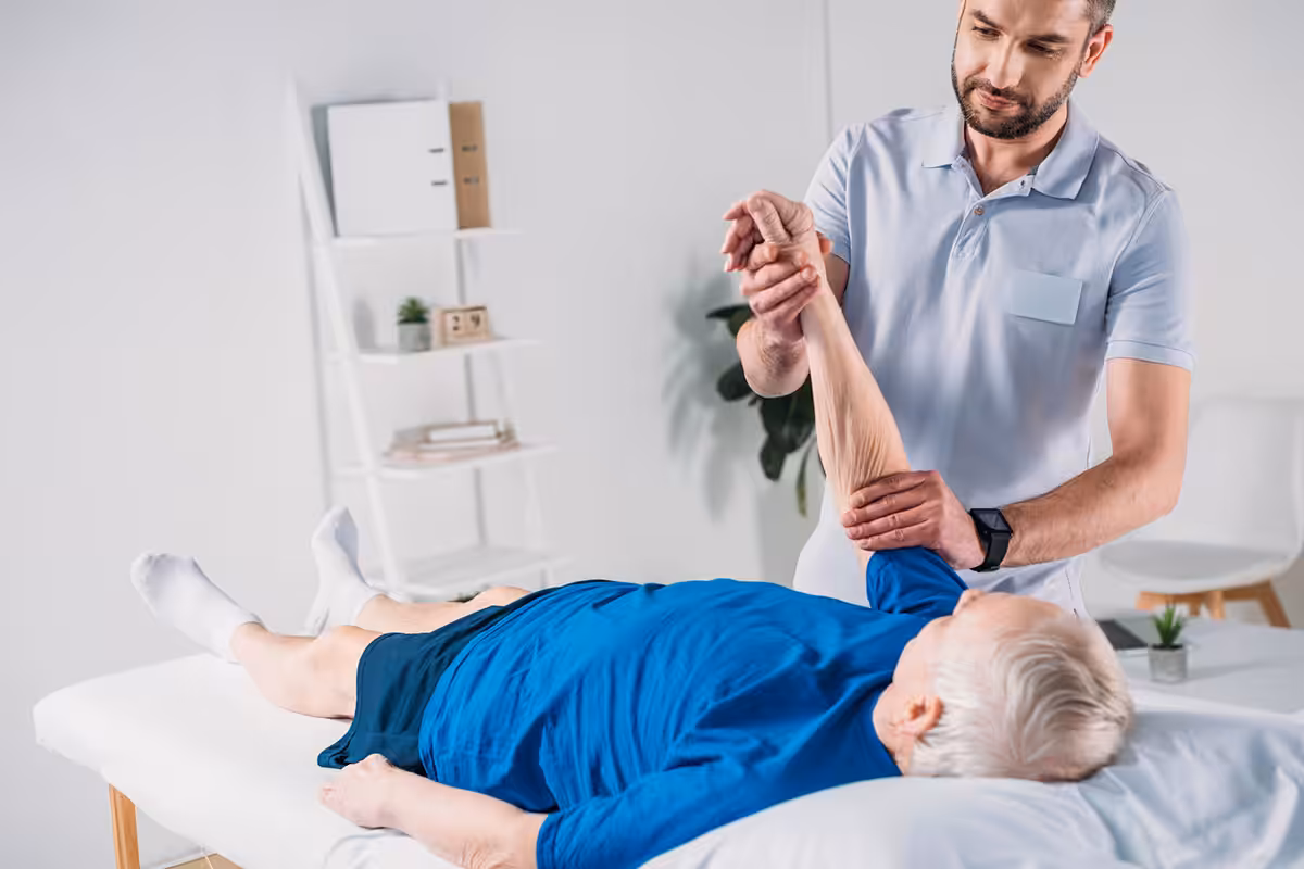 Physiotherapist performing passive arm stretch on elderly patient lying on treatment table in modern clinic
